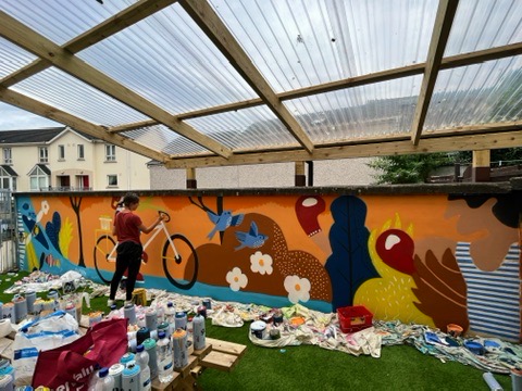 Aisling painting a bright outdoor mural with birds, bicycles and figures